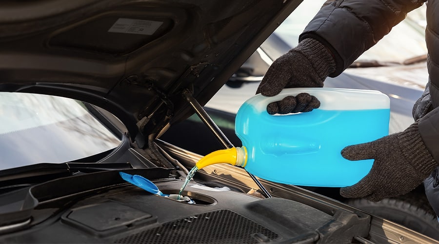Close-up detail of driver or mechanic hand in gloves pouring blue antifreeze liquid for winter car windshield screen washing with bootle watering can. Car seasonal service and maintenance.